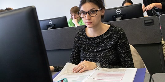 A female student at a computer with a workbook in front of her.
